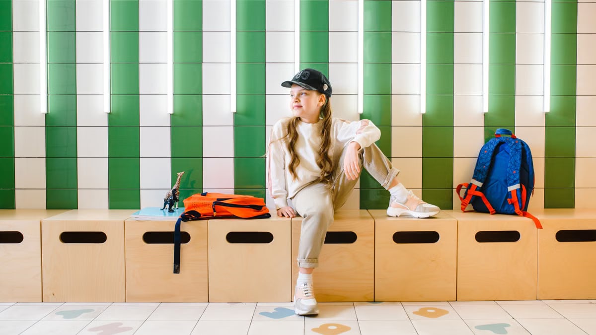 A young girl sitting indoors on a bench, with a backpack and hat, looking thoughtful.